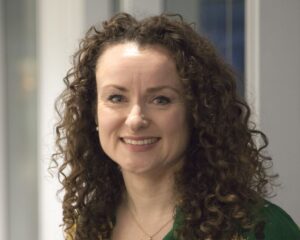 Headshot of Jane Healey Brown, with brown curly hair down to her shoulders, Jane is smiling and wearing a dark top